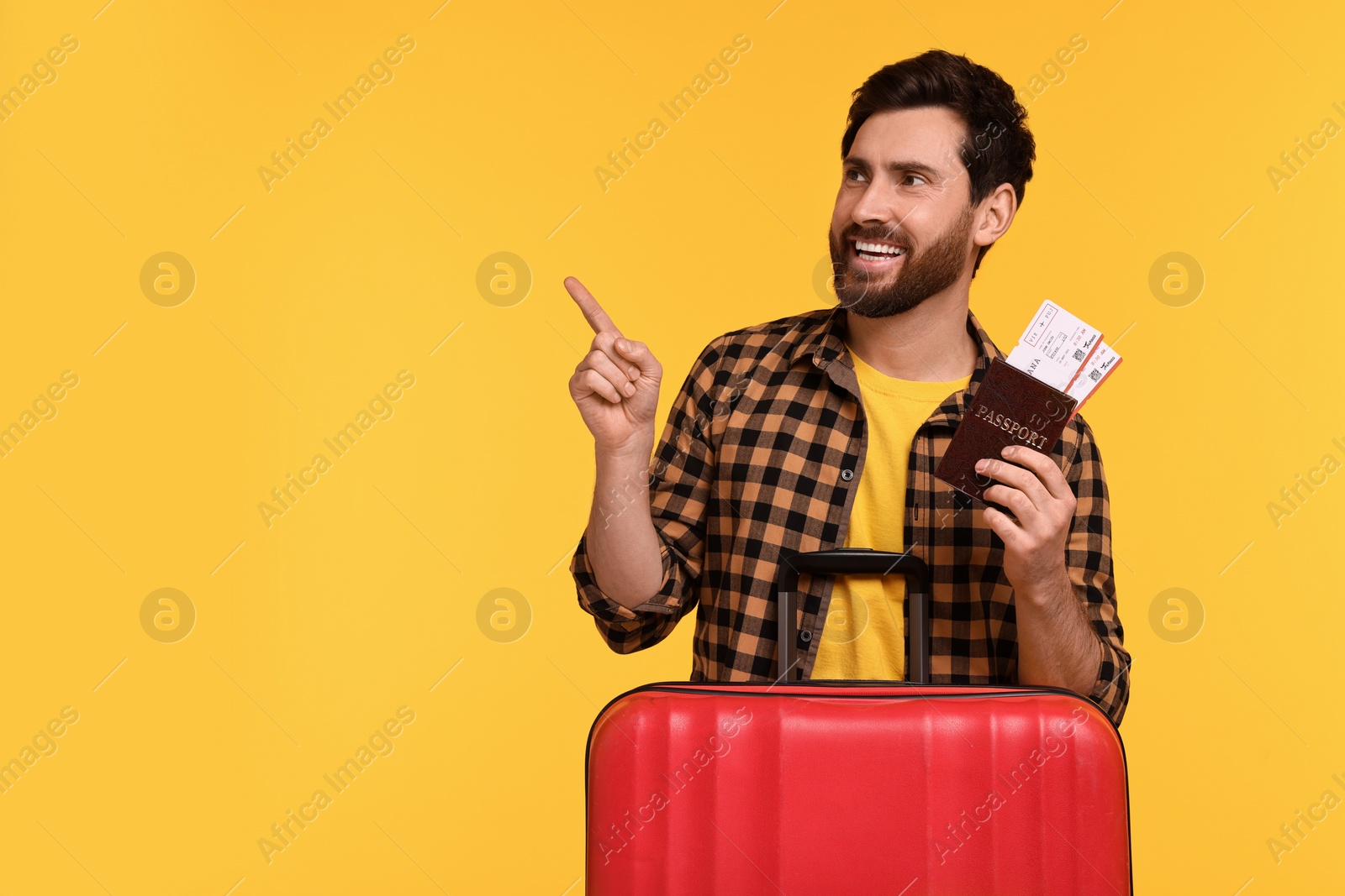 Photo of Smiling man with passport, tickets and suitcase pointing at something on yellow background. Space for text
