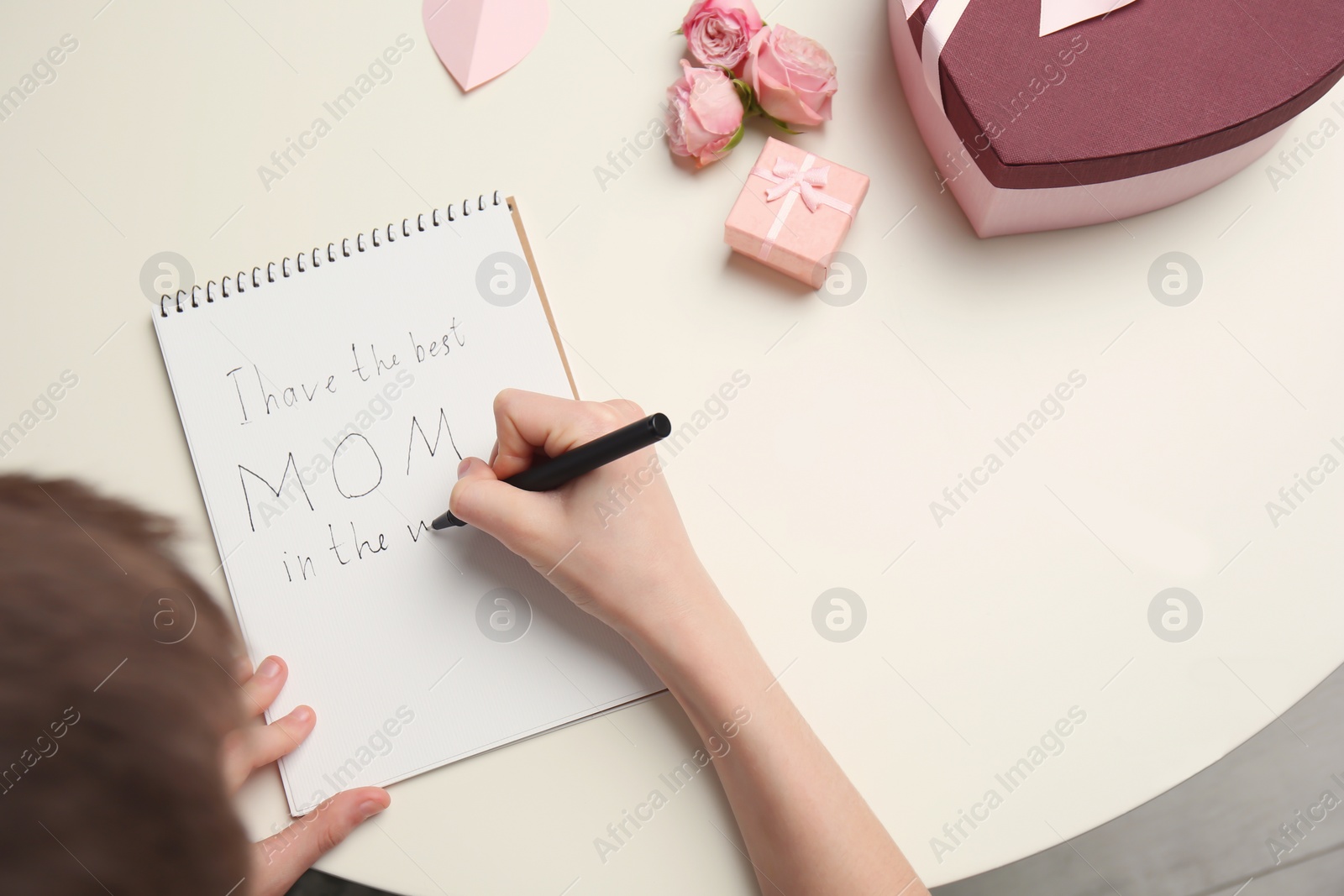 Photo of Little child writing congratulation for Mother's Day in notebook on light background, top view