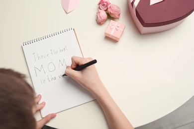 Photo of Little child writing congratulation for Mother's Day in notebook on light background, top view