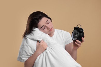 Photo of Unhappy young woman with pillow and alarm clock on beige background. Insomnia problem