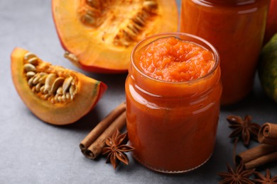 Photo of Jars of pumpkin jam, star anise, cinnamon and fresh pumpkin on grey table,closeup