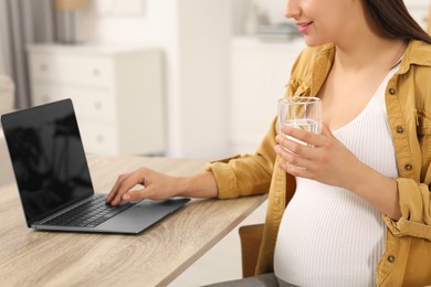 Photo of Pregnant woman with glass of water and laptop at wooden table in room, closeup