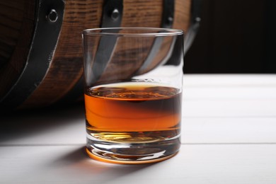 Photo of Glass of tasty whiskey and barrel on white wooden table, closeup