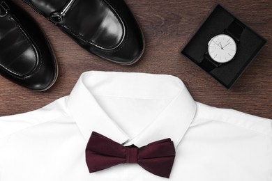 Photo of White shirt with stylish burgundy bow tie, wristwatch and shoes on wooden background, flat lay