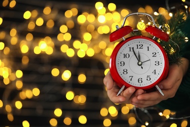 Woman holding alarm clock against blurred lights, closeup with space for text. New Year countdown