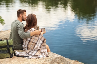 Photo of Cute couple with mugs and plaid near lake. Camping season