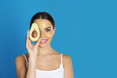 Photo of Young woman with cleansing mask on her face holding avocado against color background, space for text. Skin care