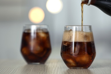 Pouring cola from bottle into glass with ice cubes on table against blurred background. Space for text