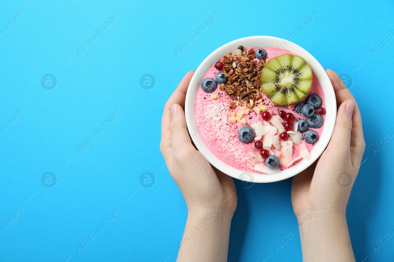 Photo of Woman holding tasty smoothie bowl with fresh kiwi fruit, berries and granola at light blue table, top view. Space for text