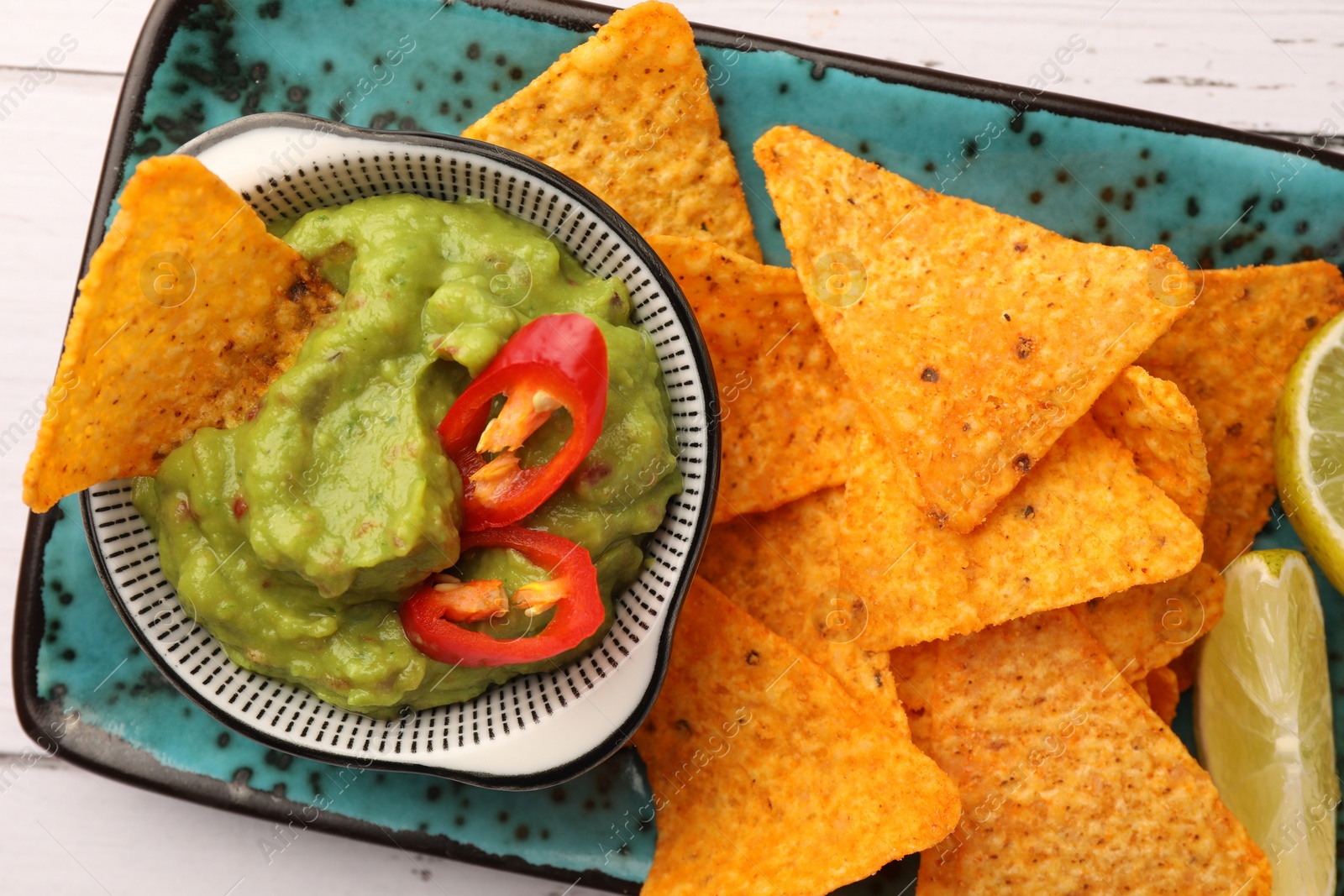 Photo of Bowl of delicious guacamole with chili pepper, nachos chips and lime on white wooden table, top view