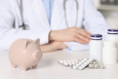 Photo of Doctor at white table indoors, focus on piggy bank and pills