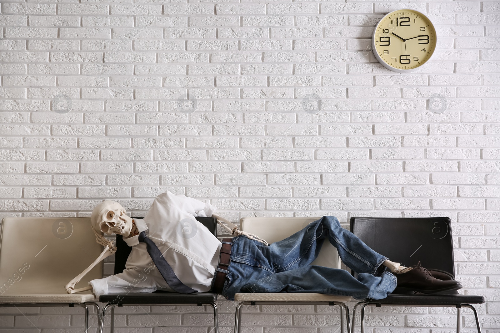 Photo of Human skeleton in office wear lying on chairs near brick wall indoors