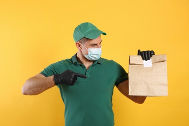 Photo of Courier in medical mask holding paper bag with takeaway food on yellow background. Delivery service during quarantine due to Covid-19 outbreak