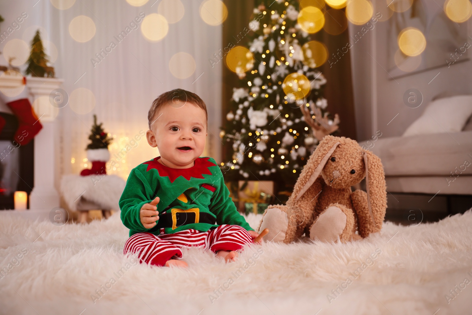 Photo of Baby wearing cute elf costume on floor in room decorated for Christmas