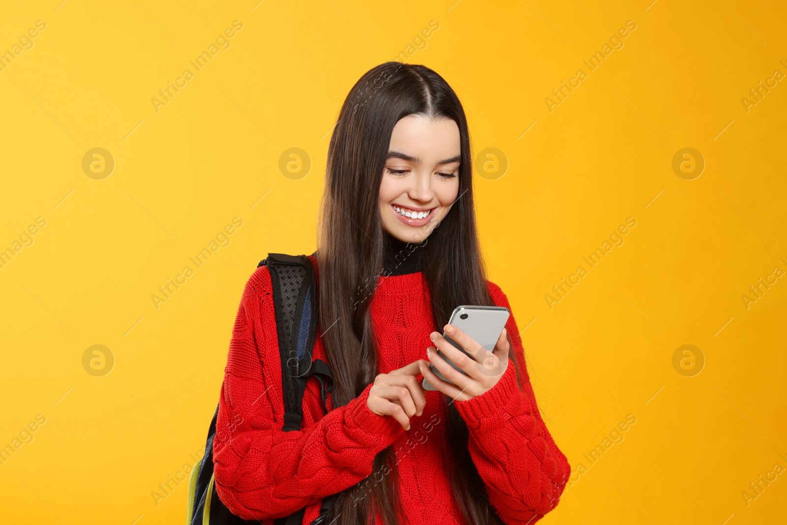 Photo of Happy teenage girl with backpack and smartphone on yellow background