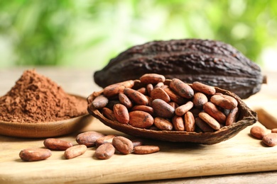 Photo of Board with cocoa pods, beans and powder on table against blurred green background