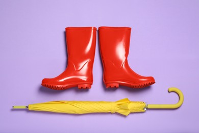 Photo of Pair of red rubber boots with yellow umbrella on violet background, flat lay