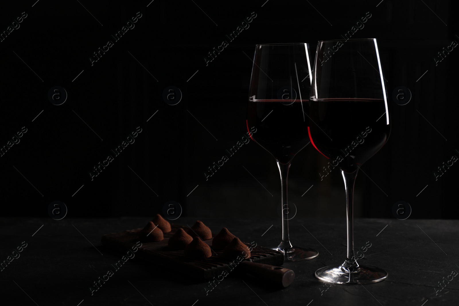 Photo of Glasses of red wine and chocolate truffles on black table in darkness, space for text