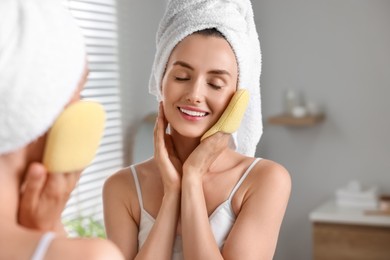 Photo of Happy young woman washing her face with sponge near mirror in bathroom