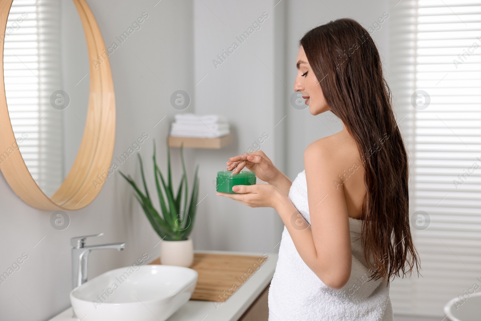 Photo of Young woman holding jar of aloe hair mask near mirror in bathroom