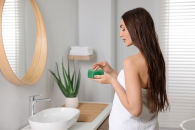 Young woman holding jar of aloe hair mask near mirror in bathroom