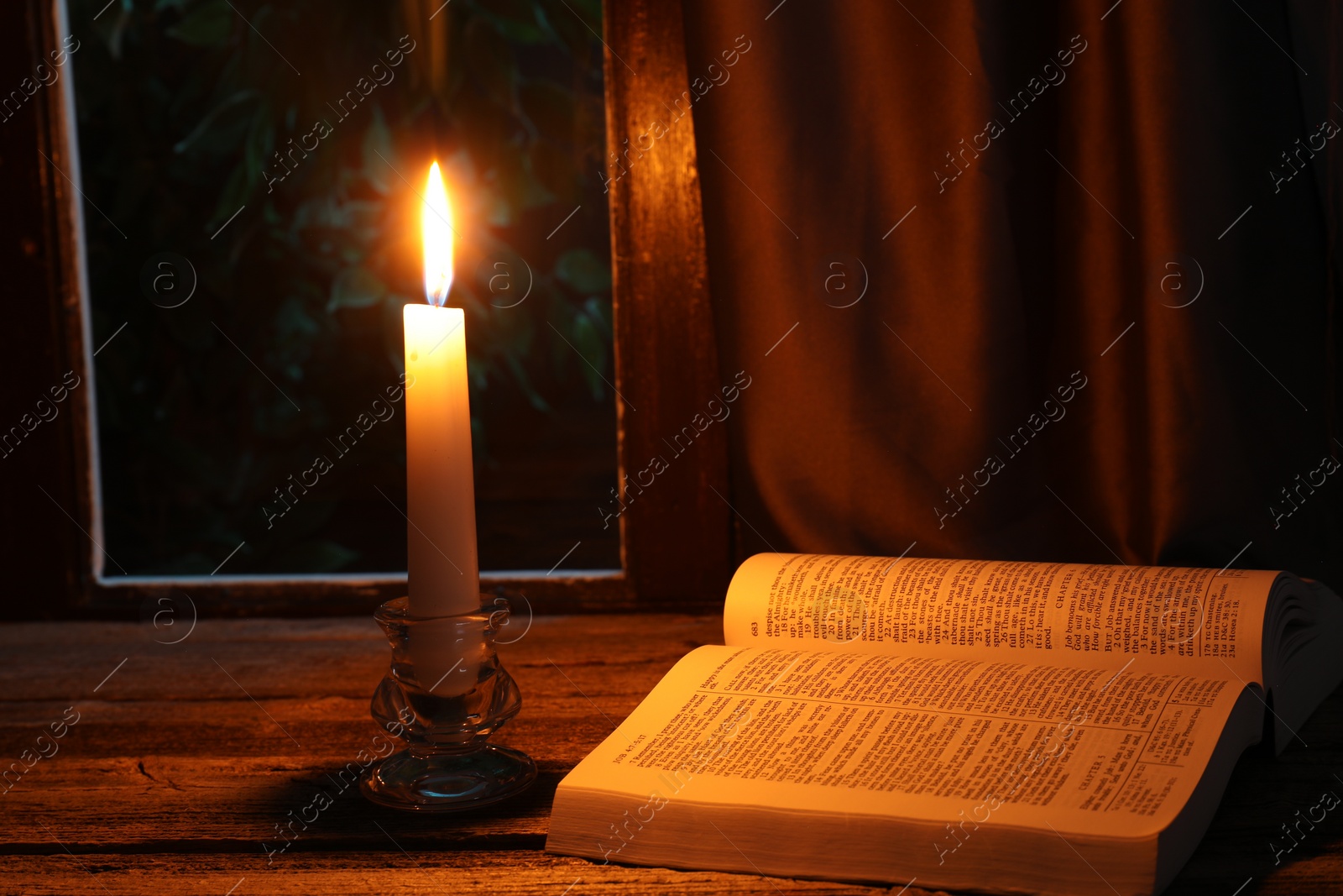 Photo of Burning candle and Bible on wooden table near window at night