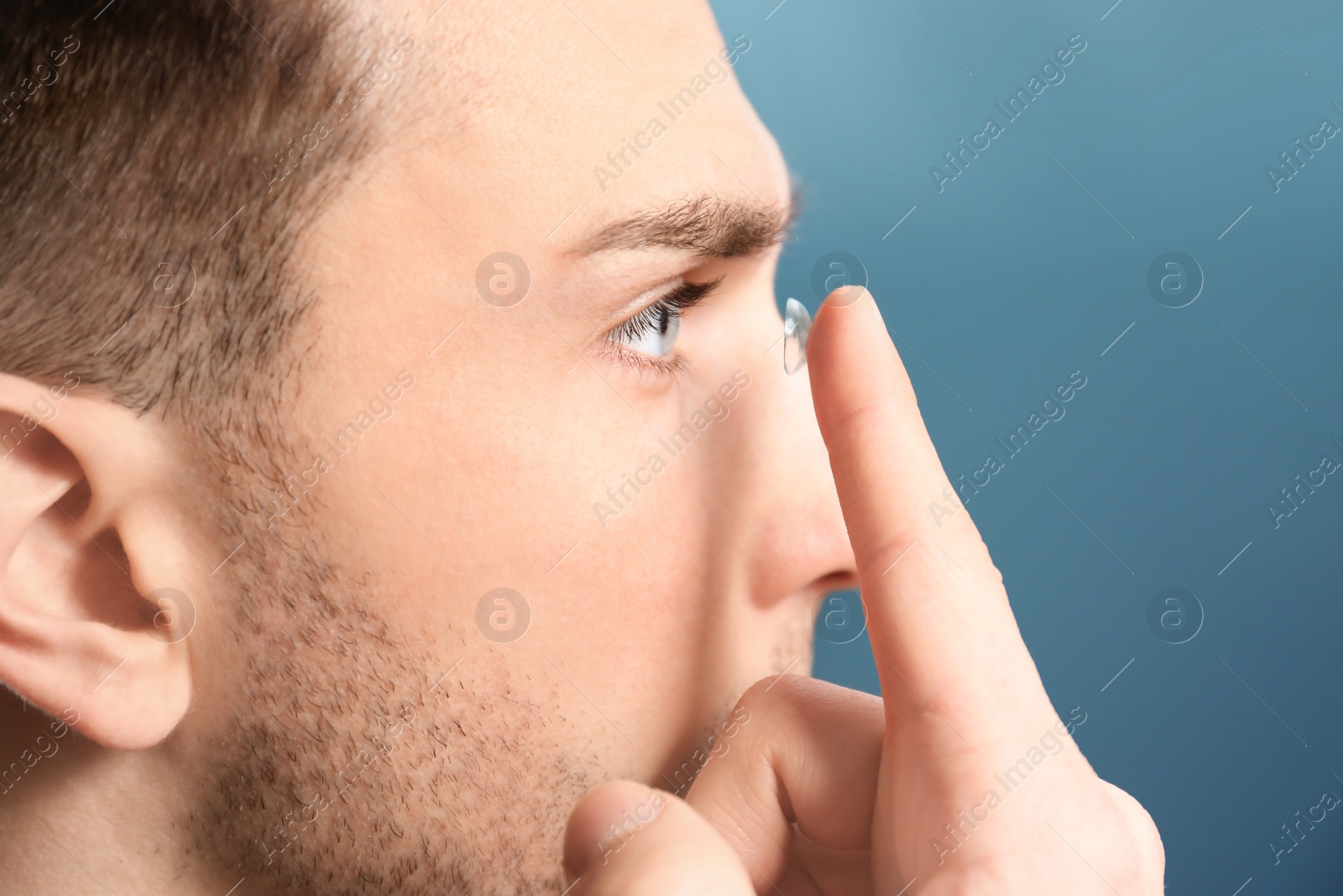 Photo of Young man putting contact lens in his eye on color background