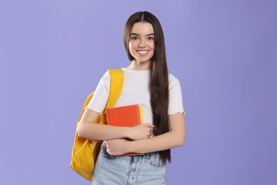 Photo of Happy teenage girl with backpack and textbooks on violet background