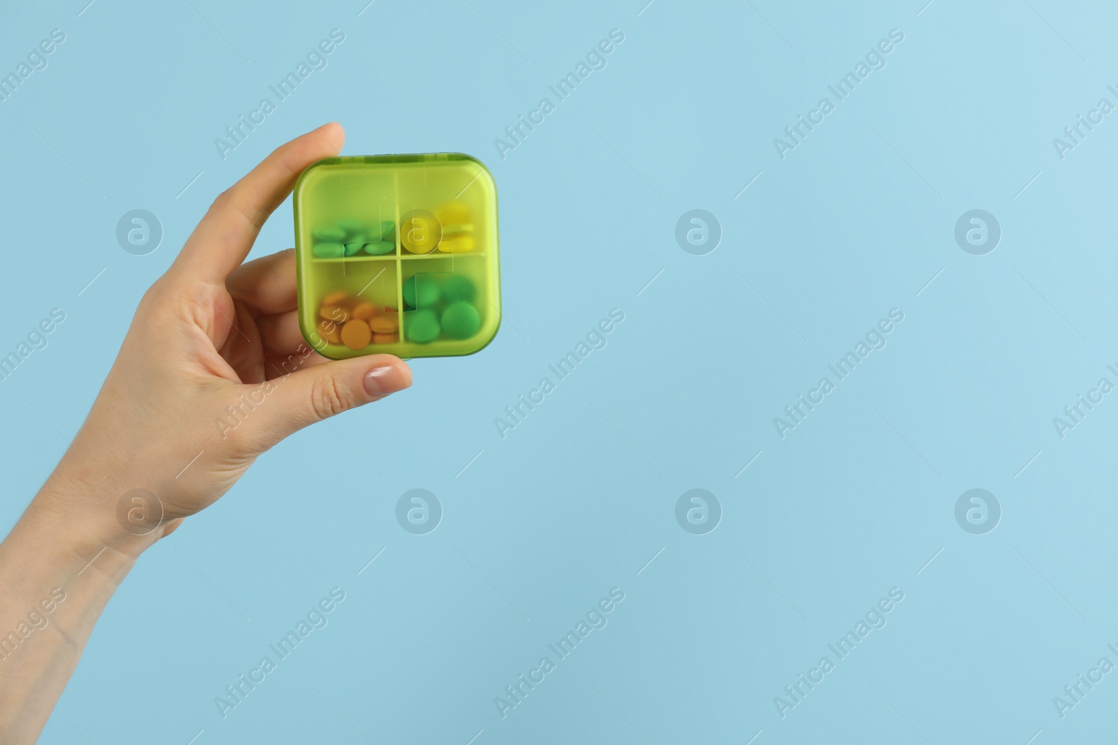 Photo of Woman holding plastic box with different pills on light blue background, closeup. Space for text