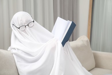 Photo of Creepy ghost. Person covered with white sheet reading book on sofa at home