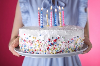 Woman holding birthday cake with burning candles on pink background, closeup