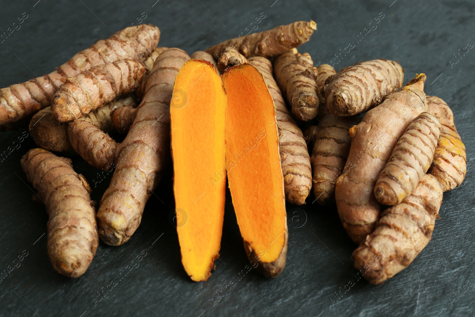 Photo of Whole and cut turmeric roots on black textured table, closeup