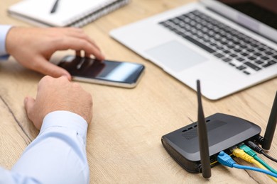 Photo of Man with laptop and smartphone connecting to internet via Wi-Fi router at wooden table indoors, closeup