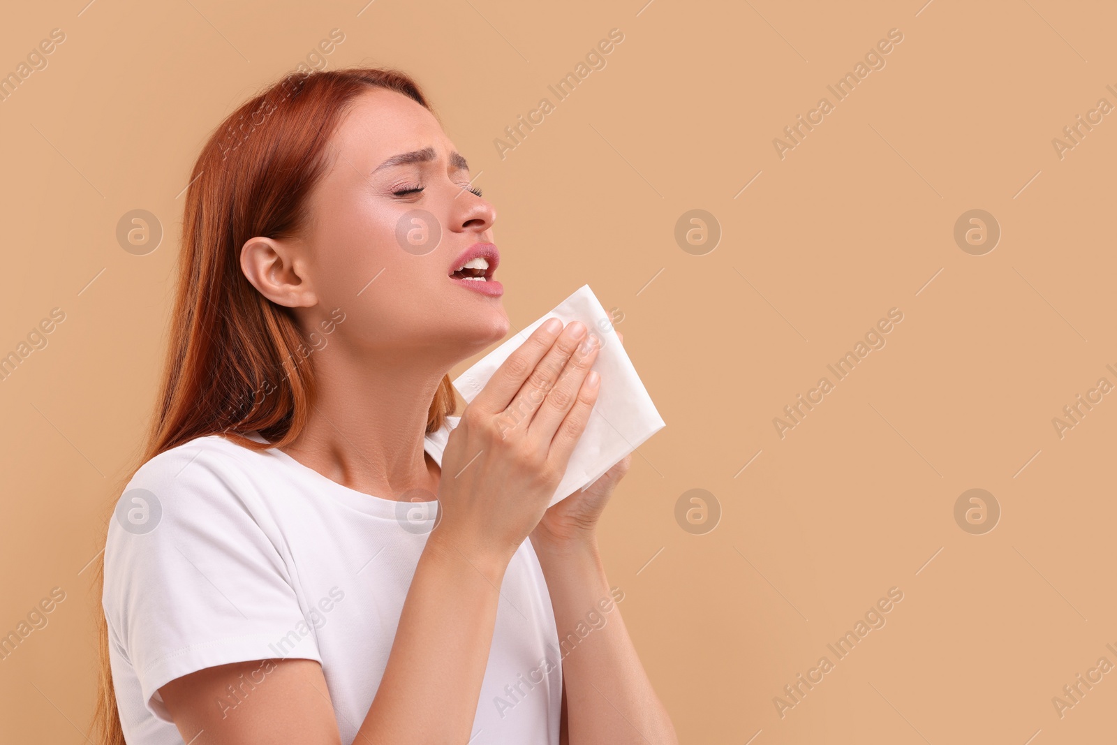 Photo of Suffering from allergy. Young woman with tissue sneezing on beige background, space for text