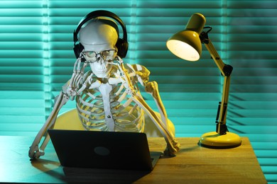 Photo of Waiting concept. Human skeleton sitting at wooden table with laptop and headphones indoors