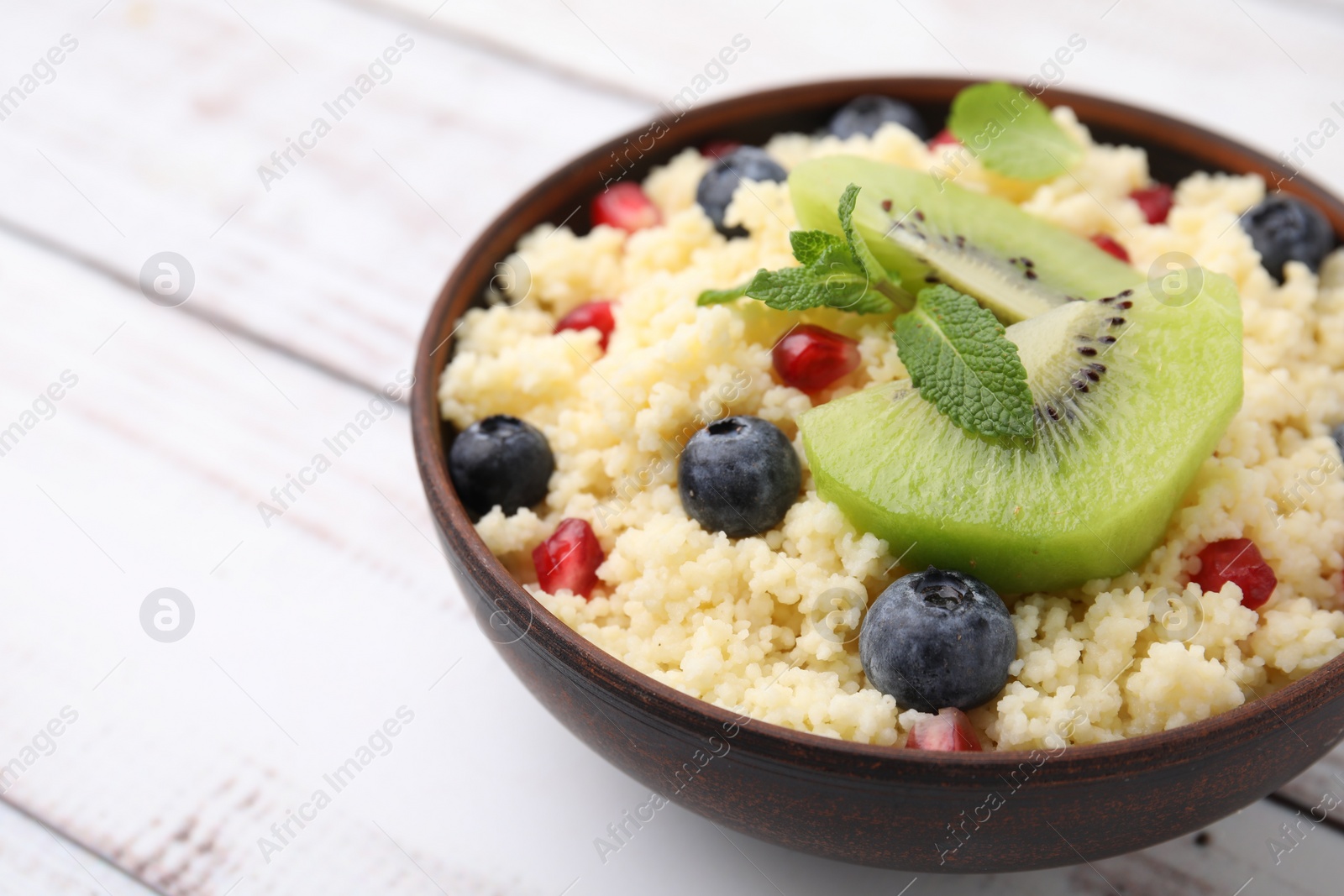 Photo of Bowl of tasty couscous with kiwi, blueberries and pomegranate on white table, closeup. Space for text