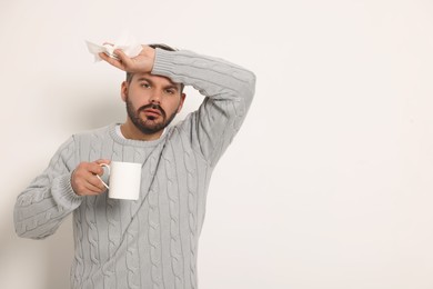 Photo of Sick man with tissue and cup of drink on light grey background, space for text. Cold symptoms