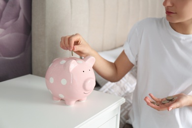 Photo of Woman putting money into piggy bank at nightstand indoors, closeup