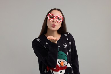 Happy young woman in Christmas sweater and funny glasses blowing kiss on grey background