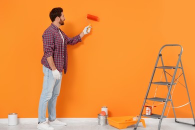 Photo of Designer painting orange wall with roller, folding ladder and cans of dye on floor indoors