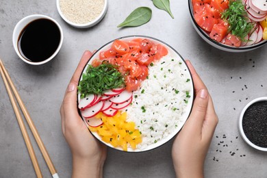 Woman holding delicious poke bowl with salmon and vegetables at light grey table, top view
