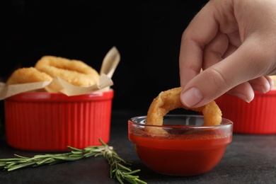 Woman dipping crunchy fried onion ring in tomato sauce on black background, closeup
