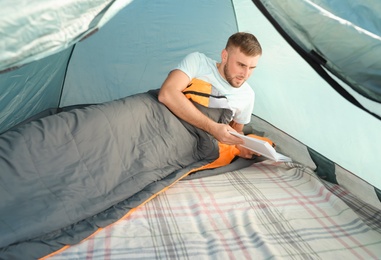 Young man reading book in sleeping bag inside of camping tent