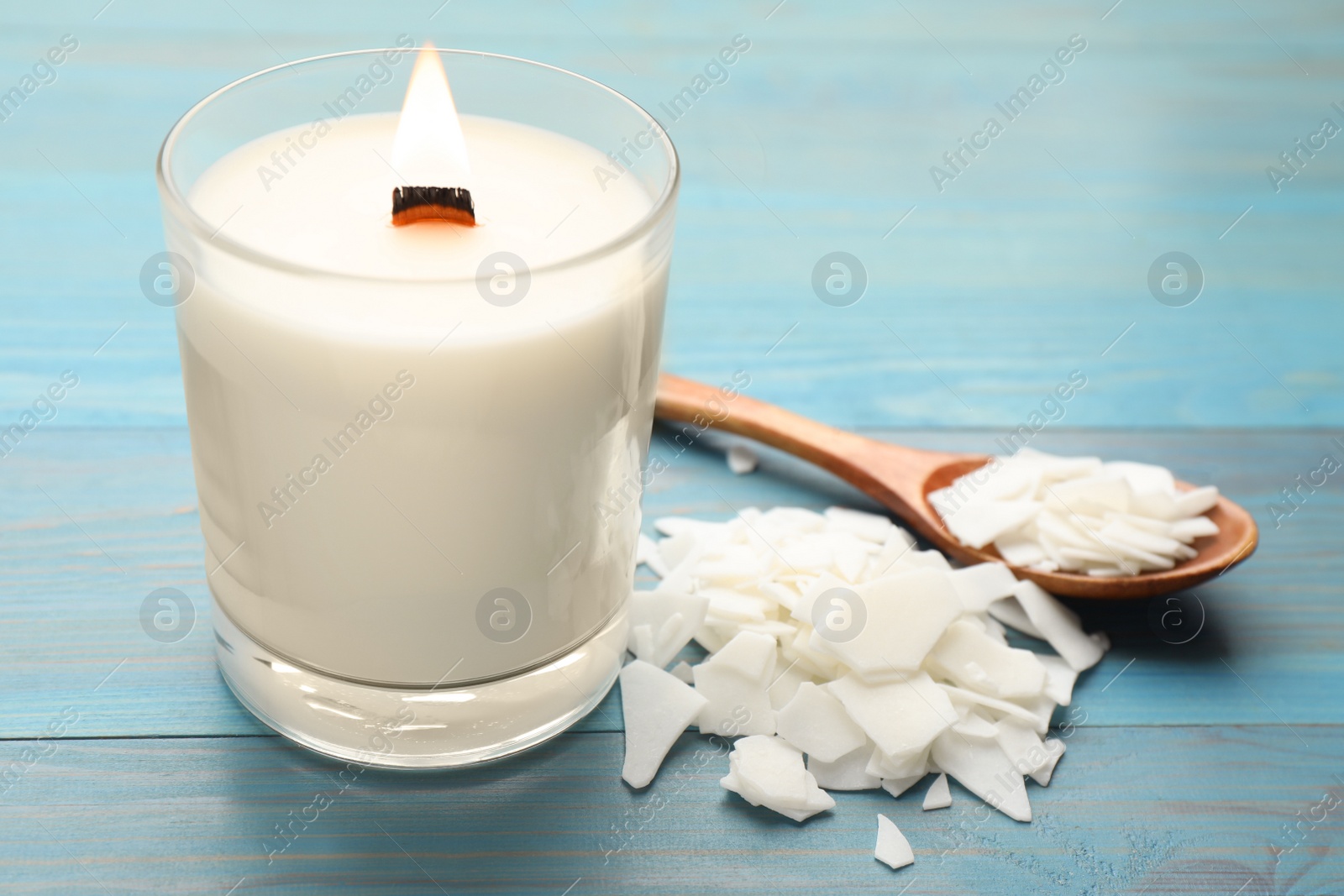 Photo of Burning soy candle, wax flakes and spoon on light blue wooden table