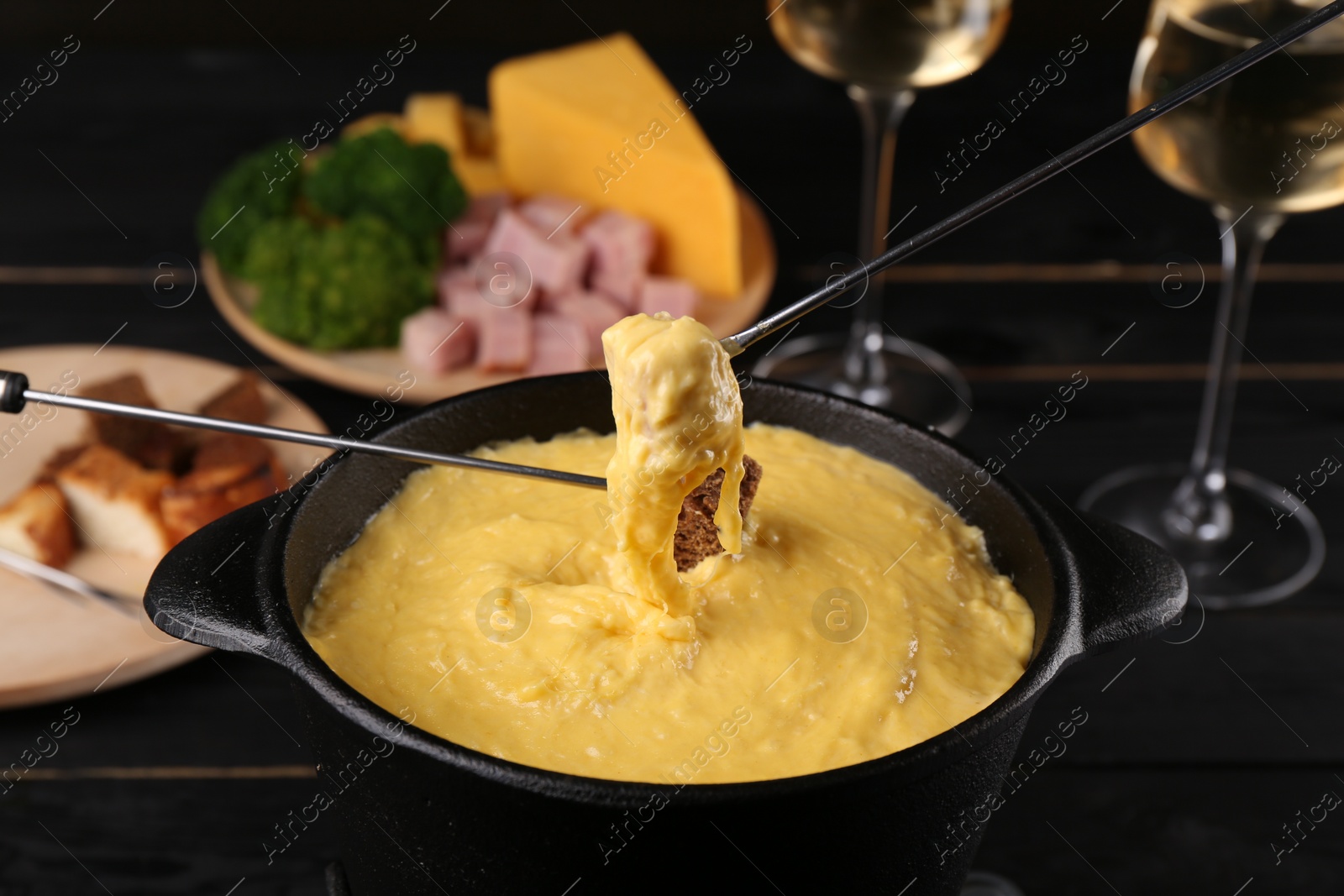 Photo of Dipping pieces of ham and bread into fondue pot with melted cheese on black wooden table, closeup