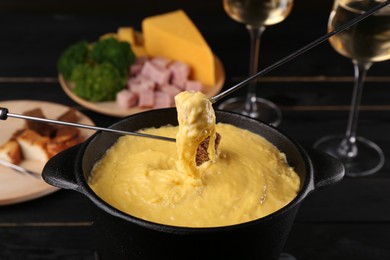 Dipping pieces of ham and bread into fondue pot with melted cheese on black wooden table, closeup