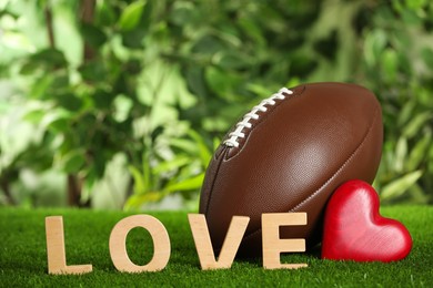 American football ball, word Love and red heart on green grass against blurred background. Space for text