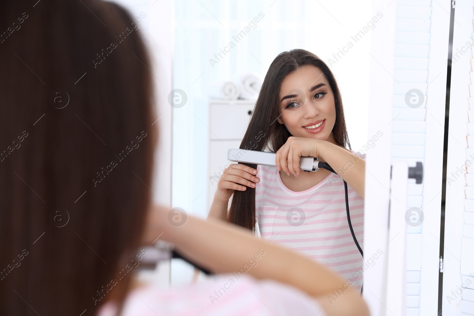 Photo of Happy woman using hair iron in front of mirror