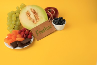 Photo of Label with word Fructose, delicious fruits, berries and pieces of chocolate on yellow background, space for text