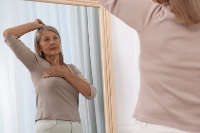 Photo of Beautiful senior woman doing breast self-examination near mirror indoors